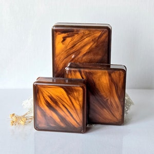 May include: Three square boxes in varying sizes, featuring a marbled pattern in shades of brown and amber. The boxes have rounded corners and a glossy finish, suggesting a vintage or decorative item. The boxes are arranged together, showcasing their different sizes.