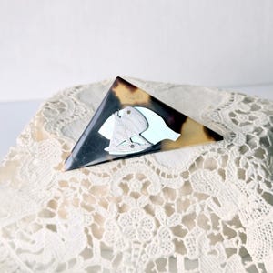 May include: A vintage triangular brooch with a tortoiseshell pattern and a light blue and white design. The brooch is set on a white lace surface. The brooch features a stylized design.