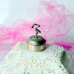 May include: A small, silver-toned trinket box with a decorative stork figurine on the lid. The box sits on a white lace doily, with a sheer pink fabric in the background. The box is round and has a detailed border.