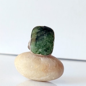 May include: A silver ring featuring a large, rectangular green stone with dark green and black marbling. The ring is set on a light-colored, oval stone against a white background. The gemstone has a polished surface.