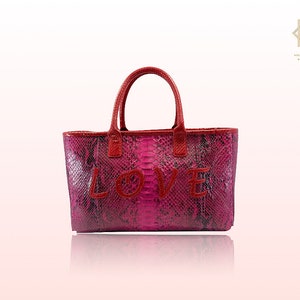 Peut inclure: Un sac cabas rose et rouge avec un motif peau de serpent et le mot "LOVE" brodé en rouge.