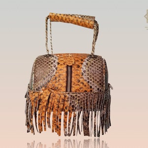 Peut inclure: Un petit sac à main en cuir de serpent brun, orange et gris avec une bordure en franges. Le sac a une poignée supérieure et une fermeture à glissière.