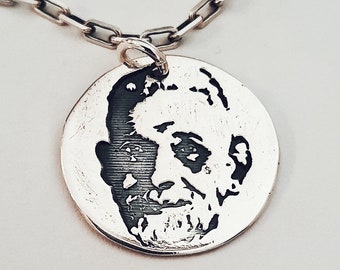 May include: A silver pendant necklace featuring a circular pendant with a detailed engraved portrait of a person. The pendant hangs from a silver chain with a link design. The portrait is in black and white.