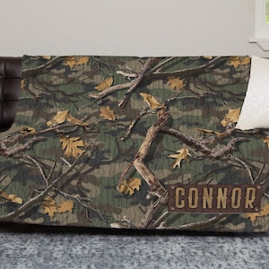 May include: A camouflage-patterned throw blanket with a brown rectangular nameplate that reads "CONNOR". The blanket features a design of tree branches and leaves in shades of green, brown, and tan. The blanket is draped over a dark brown leather couch.