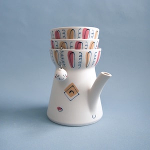 Può includere: Una teiera in ceramica bianca con beccuccio e tre tazze impilate. Le tazze e la teiera sono decorate con disegni astratti rosa, gialli e neri. Una piccola palla bianca sorridente con una catena pende dalla teiera.