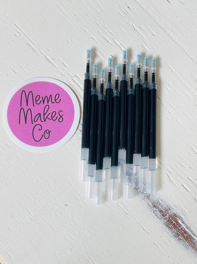 May include: A pack of 12 black ink refills for gel pens. The refills have clear plastic barrels and blue tips. The refills are arranged in a stack on a white wooden surface. The text "Meme Makes Co." is visible on a pink sticker in the background.
