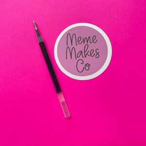 May include: A black pen with a clear plastic barrel on a pink background. A sticker with the text "Meme Makes Co." is in the upper right corner of the image.