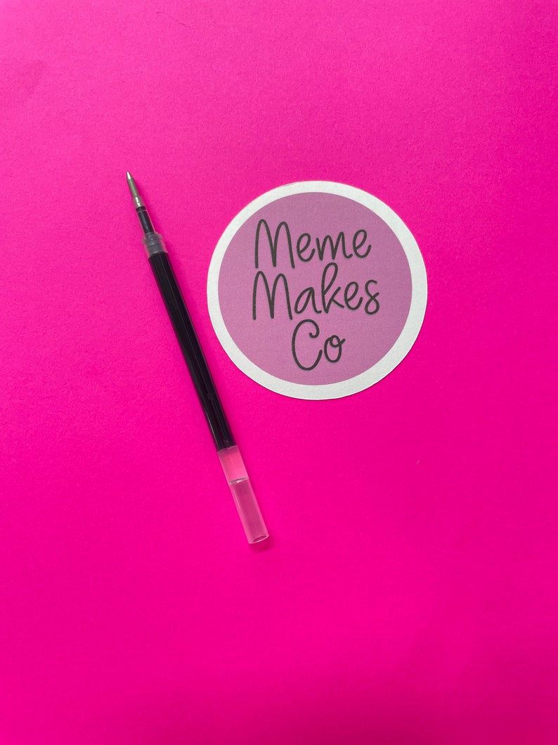 May include: A black pen with a clear plastic barrel on a pink background. A sticker with the text "Meme Makes Co." is in the upper right corner of the image.