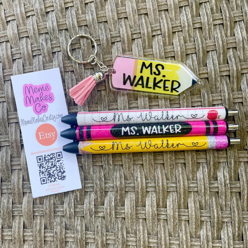 以下が含まれることがあります： 「Ms. Walker」と名前が入ったパーソナライズされたペンが3本。ペンはキラキラと輝き、ピンク、イエロー、ホワイトのカラーリングが施されています。ペンは、ピンクのタッセルが付いたキーホルダーと、透明なアクリル製の鉛筆チャーム（「MS. WALKER」の文字入り）の隣に置かれています。