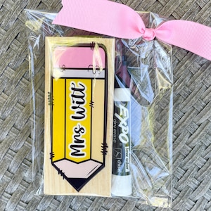 May include: A clear cellophane bag tied with a pink ribbon contains a wooden pencil-shaped bookmark and a white dry-erase marker. The bookmark has the text "Mrs. Witt" on it.