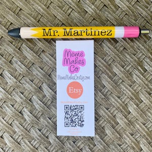 以下が含まれることがあります： ピンクの文字で「Meme Makes Co」と書かれた白い名刺。名刺には「MemeMakesCoetsy.com」というウェブサイト、赤い円の中に「Etsy」という単語、QRコードも記載されています。