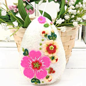 Könnte beinhalten: Weißes Osterei-Ornament aus Filz mit rosa und roten Blumenstickereien. Der eiförmige Schmuck hat eine Schnur zum Aufhängen und ist vor einem Korb mit rosa, weißen und grünen Blumen platziert.