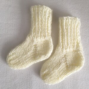 May include: A pair of ivory-colored, knitted baby socks. The socks are made of a soft yarn and have a ribbed cuff and a textured knit pattern. The socks are small, suggesting they are for a newborn or infant.