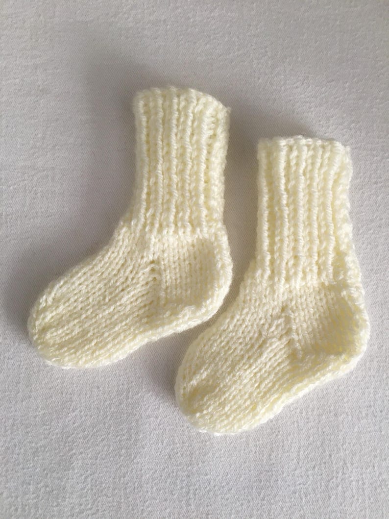 May include: A pair of ivory-colored, knitted baby socks. The socks are made of a soft yarn and have a ribbed cuff and a textured knit pattern. The socks are small, suggesting they are for a newborn or infant.