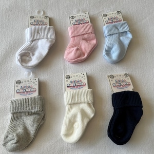 May include: Six pairs of baby socks in different colors, including white, pink, blue, gray, and black. The socks are all folded and displayed on a white surface. Each pair has a small tag that says "Baby Socks Cotton Socks".