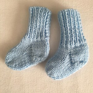 May include: A pair of light blue knitted baby socks. The socks are made of soft yarn and have a ribbed cuff and a textured stitch pattern. The socks are designed for infants.