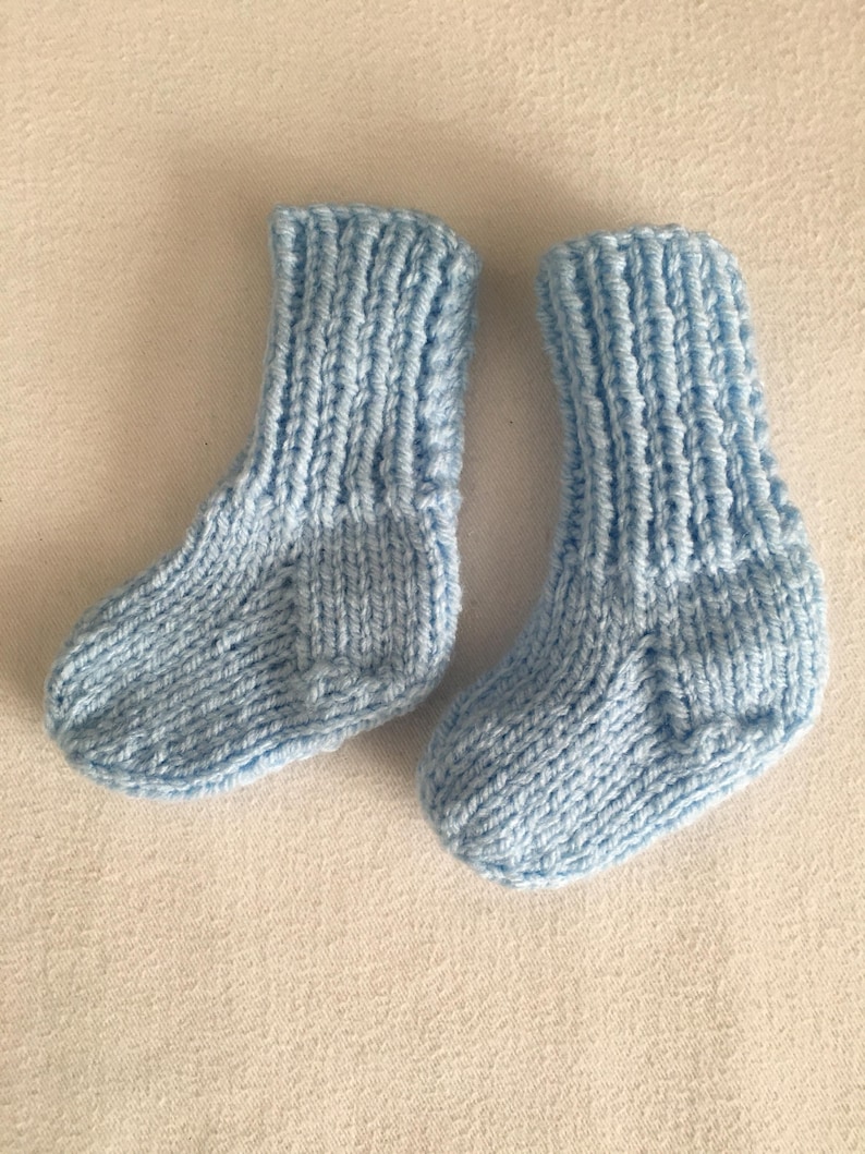 May include: A pair of light blue knitted baby socks. The socks are made of soft yarn and have a ribbed cuff and a textured stitch pattern. The socks are designed for infants.