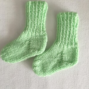 May include: A pair of hand-knitted baby socks in a soft, light green color. The socks are made with a ribbed cuff and a simple knit stitch, suggesting a cozy and comfortable design. The socks are likely made of a soft yarn, perfect for a baby.