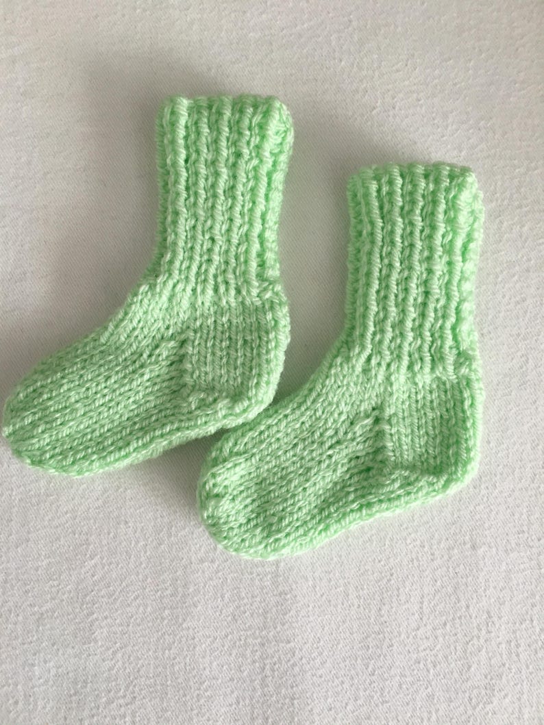 May include: A pair of hand-knitted baby socks in a soft, light green color. The socks are made with a ribbed cuff and a simple knit stitch, suggesting a cozy and comfortable design. The socks are likely made of a soft yarn, perfect for a baby.
