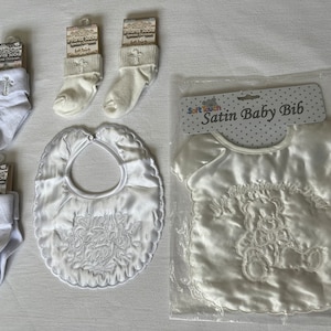 May include: White satin baby bib with embroidered cross and the text "My First Holy Communion Day".  The bib is packaged in a clear plastic bag with a white label that reads "Soft Touch Satin Baby Bib".  The image also includes four pairs of white socks with embroidered crosses.