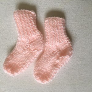 May include: A pair of soft, light pink knitted baby socks. The socks are made of a textured yarn and are designed to keep little feet warm. The socks are displayed on a white surface.