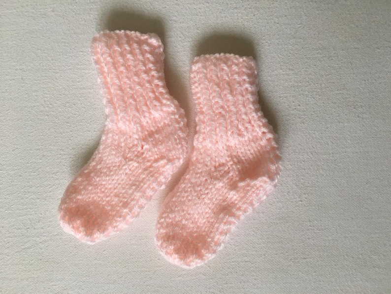 May include: A pair of soft, light pink knitted baby socks. The socks are made of a textured yarn and are designed to keep little feet warm. The socks are displayed on a white surface.