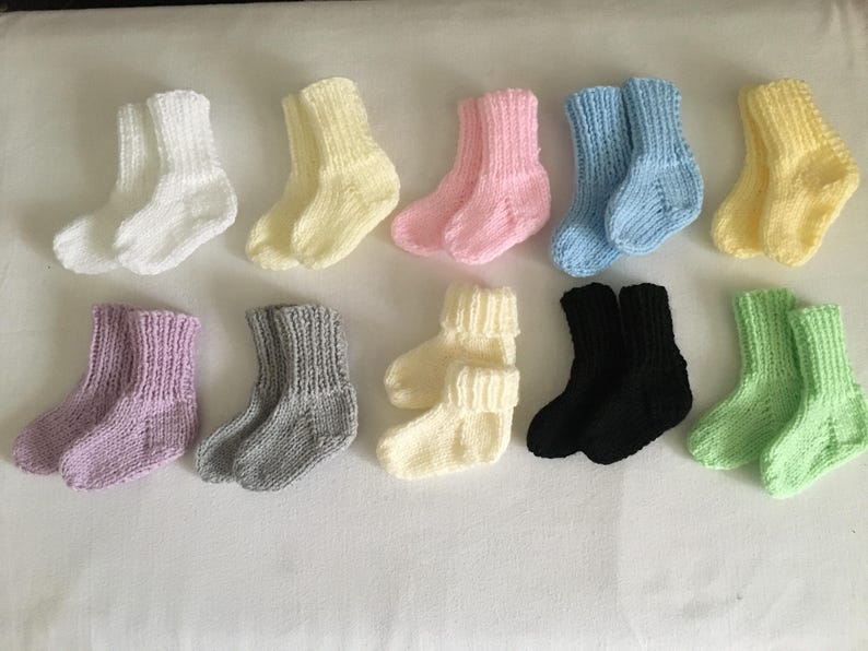 May include: A collection of ten pairs of knitted baby socks in various colors, including white, yellow, pink, light blue, lavender, gray, cream, black, and light green. The socks are arranged on a white surface.
