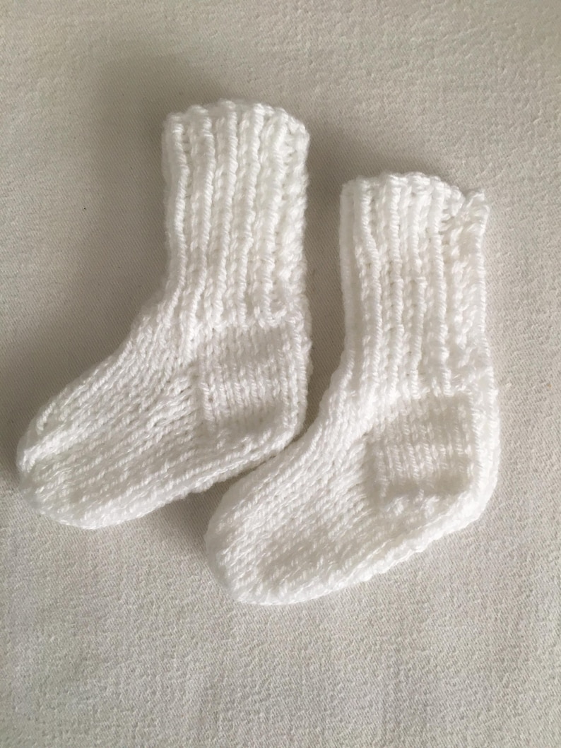 May include: A pair of small, knitted white socks. The socks are made of a soft, textured yarn and have a ribbed cuff. The socks are designed for a baby or small child.