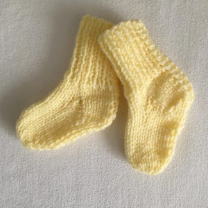 May include: A pair of soft, yellow knitted baby socks. The socks are made of a textured yarn and have a ribbed cuff. The socks are small, designed for a newborn or infant. The socks are a pale yellow color.