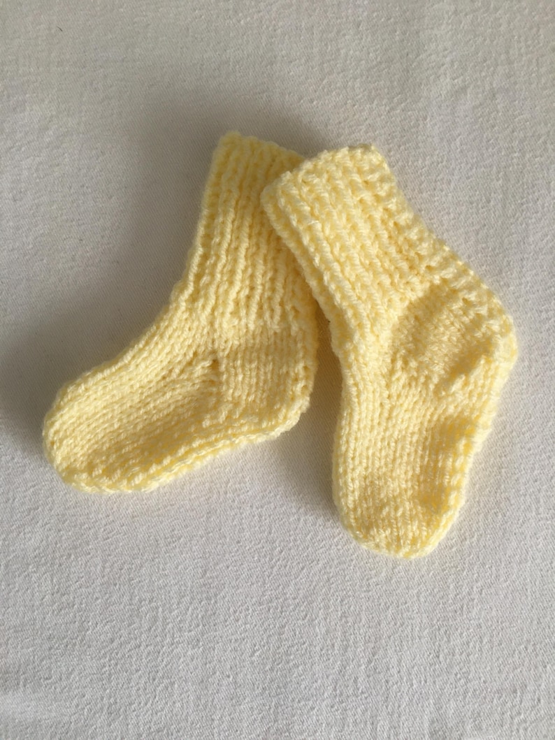 May include: A pair of soft, yellow knitted baby socks. The socks are made of a textured yarn and have a ribbed cuff. The socks are small, designed for a newborn or infant. The socks are a pale yellow color.