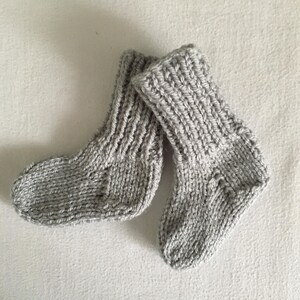 May include: A pair of hand-knitted baby socks in a light gray color. The socks are made with a thick yarn, featuring a ribbed texture on the cuffs and a simple knit pattern throughout the body. The socks are designed to keep a baby's feet warm.
