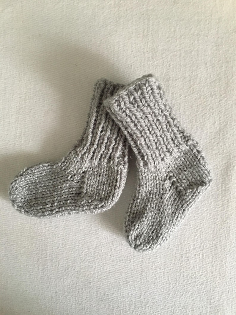 May include: A pair of hand-knitted baby socks in a light gray color. The socks are made with a thick yarn, featuring a ribbed texture on the cuffs and a simple knit pattern throughout the body. The socks are designed to keep a baby's feet warm.
