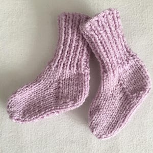 May include: A pair of knitted baby socks in a soft lavender color. The socks are made with a ribbed knit pattern and are designed for small feet. The texture of the yarn is visible, suggesting a handmade item.