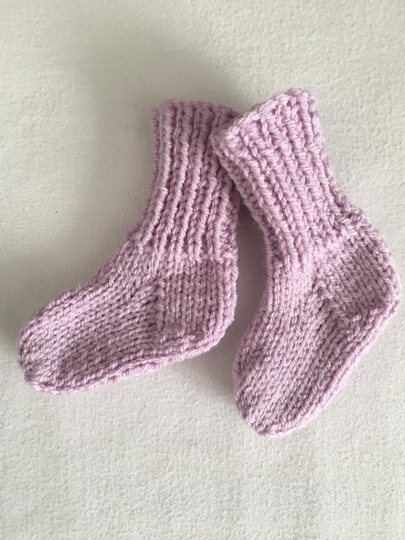 May include: A pair of knitted baby socks in a soft lavender color. The socks are made with a ribbed knit pattern and are designed for small feet. The texture of the yarn is visible, suggesting a handmade item.