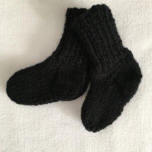 May include: A pair of black knitted baby socks. The socks are made of a thick yarn and have a ribbed texture. The socks are lying on a white surface.