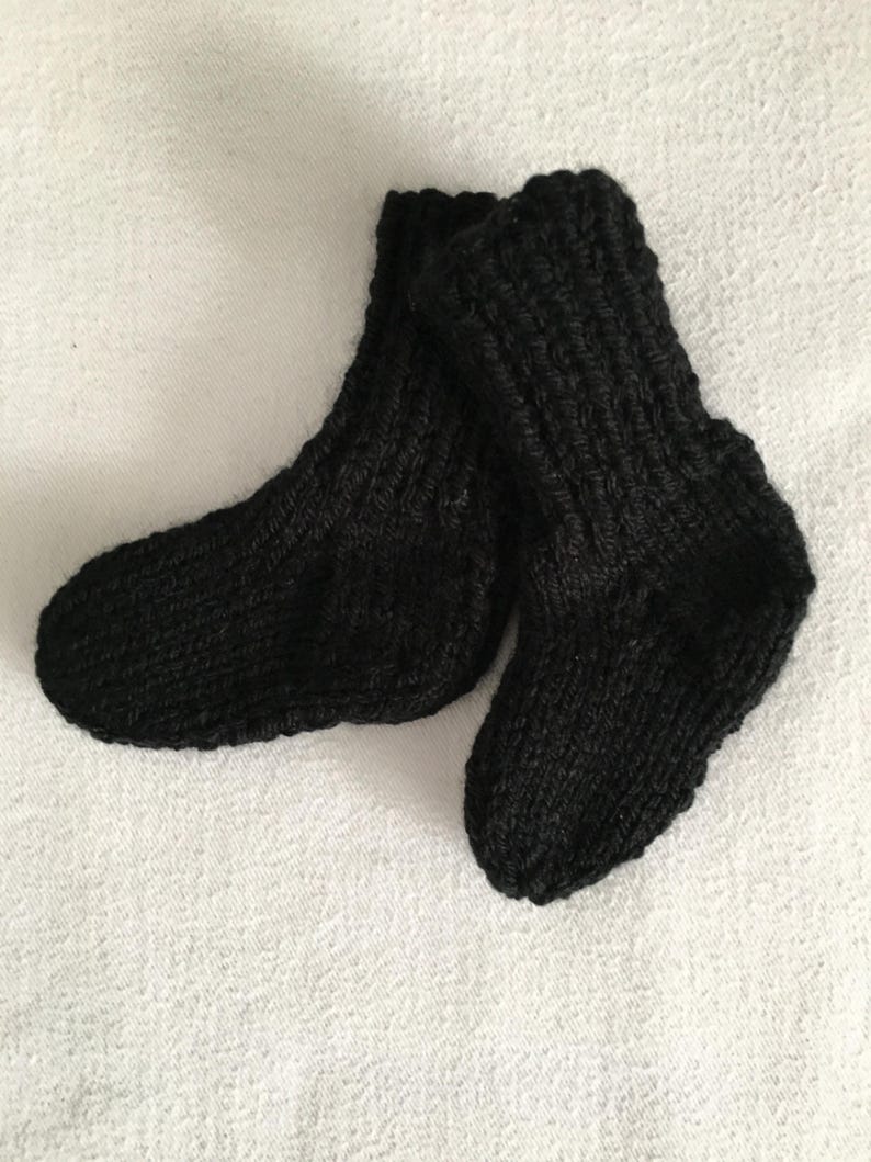 May include: A pair of black knitted baby socks. The socks are made of a thick yarn and have a ribbed texture. The socks are lying on a white surface.