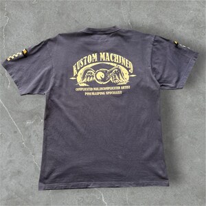 May include: Dark gray t-shirt with yellow text and graphics. The back of the shirt features the words "KUSTOM MACHINED" above an emblem with wings and a circle. Below the emblem are the words "COMPLICATED MAN, UNCOMPLICATED ARTIST PINSTRIPING SPECIALIST."