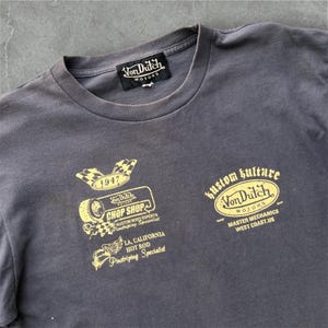 May include: Charcoal gray t-shirt with multiple gold-colored graphics. The graphics include the text "Von Dutch Motors," "Chop Shop," and "Kustom Kulture." Additional text includes "LA, California Hot Rod Pinstriping Specialist" and "Master Mechanics West Coast.US."