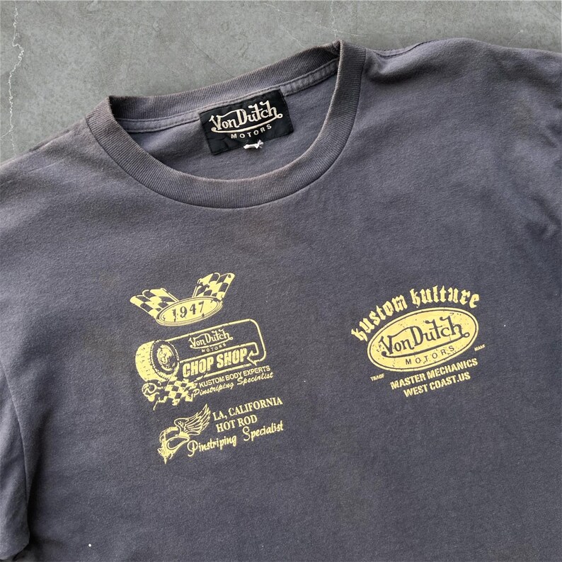 May include: Charcoal gray t-shirt with multiple gold-colored graphics. The graphics include the text "Von Dutch Motors," "Chop Shop," and "Kustom Kulture." Additional text includes "LA, California Hot Rod Pinstriping Specialist" and "Master Mechanics West Coast.US."