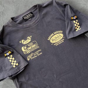 May include: Dark gray t-shirt with multiple gold-colored graphics and text. The shirt features the "Von Dutch Motors" logo, "Kustom Kulture" text, and patches on the sleeves. The graphics include checkered flags and an eye with wings.