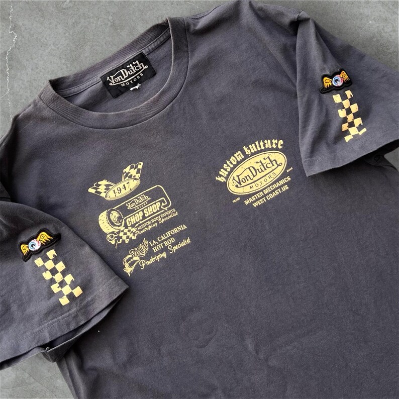 May include: Dark gray t-shirt with multiple gold-colored graphics and text. The shirt features the "Von Dutch Motors" logo, "Kustom Kulture" text, and patches on the sleeves. The graphics include checkered flags and an eye with wings.