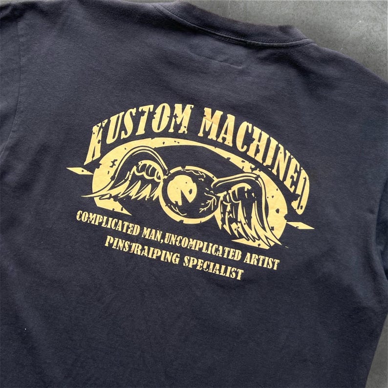 May include: Dark navy blue t-shirt with a distressed gold graphic on the back. The graphic reads "KUSTOM MACHINED" above a winged emblem, with text below that reads "COMPLICATED MAN, UNCOMPLICATED ARTIST, PINSTRIPING SPECIALIST."