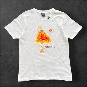 Puede incluir: Una camiseta blanca de manga corta con una ilustración de dibujos animados de un personaje de porción de pizza que lleva un gorro de chef y sostiene una pequeña caja de pizza. El gráfico incluye el texto PIZZA-LA en negro con un logo cuadrado rojo.