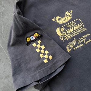 May include: Dark gray t-shirt with gold graphics and text. The sleeve features a patch with an eye and wings, and a checkered pattern. The shirt's design includes the words "Von Dutch Motors Chop Shop" and "1947".