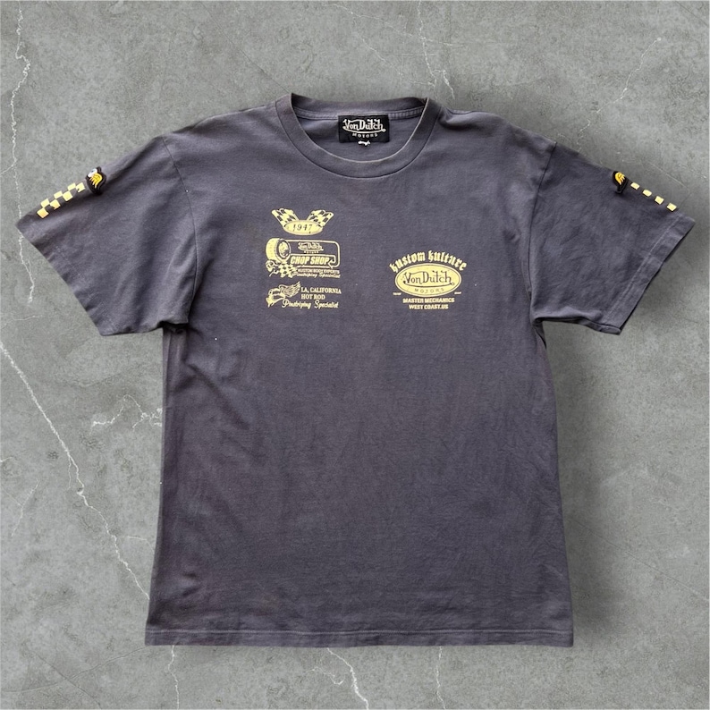 May include: Dark grey t-shirt with yellow graphics. The shirt features the text "Von Dutch" and various vintage-style logos and text, including "Kustom Kulture" and "Crop Shop". The sleeves have yellow checkered flag designs.