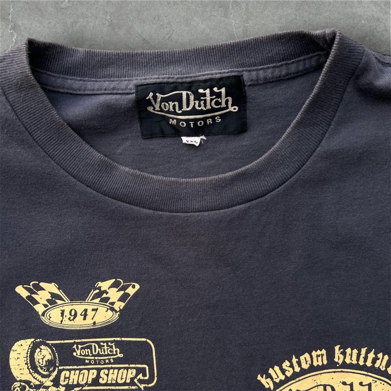 May include: Dark gray t-shirt with a crew neck and short sleeves. The shirt features a black label with "Von Dutch Motors" in gold lettering. Below, there are gold graphics including a checkered flag, the year "1947", and "Chop Shop".