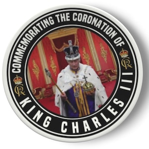 Può includere: Un bottone rotondo bianco con un bordo nero e il testo "Commemorating the Coronation of King Charles III" in nero. Il centro del bottone presenta una foto di re Carlo III con i suoi abiti da incoronazione, che tiene uno scettro e un globo.