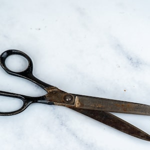 Puede incluir: Un par de tijeras vintage negras con una cuchilla oxidada y un remache plateado sobre una superficie de mármol blanco.