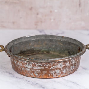 May include: An antique copper serving bowl with two ornate brass handles. The bowl has a worn, aged patina and is slightly oval in shape.