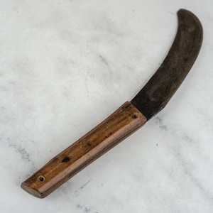 Puede incluir: Un cuchillo de hoja curva con mango de madera. La hoja es oscura y parece ser de metal. El mango es de color marrón claro. El cuchillo probablemente se utiliza para cortar o cosechar.